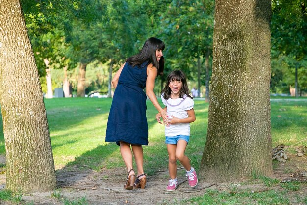 Opgewonden gelukkig meisje actieve spelletjes spelen met haar moeder buitenshuis, staande door bomen in het park en lachen. Volledige lengte. Familie buitenactiviteiten en vrijetijdsconcept