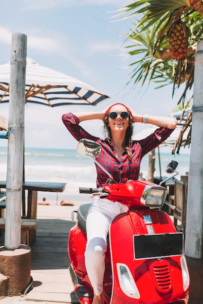 Opgewonden gelukkig stijlvol meisje draagt zwarte zonnebril en rode pet, geruite overhemd heeft plezier op rode fiets bij de oceaan en palmbladeren, zomervakantie, reizen, eiland