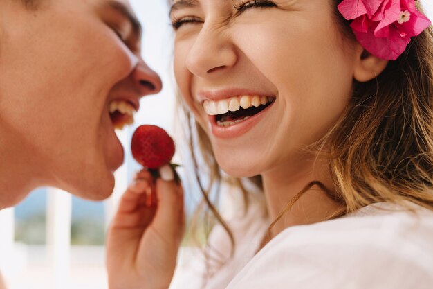 Opgewonden gelukkige jonge vrouw met schattige roze bloem in lichtbruin haar die haar lachende echtgenoot voedt met verse aardbeien. Close-upportret van romantisch genieten van huwelijksreis en bessen eten