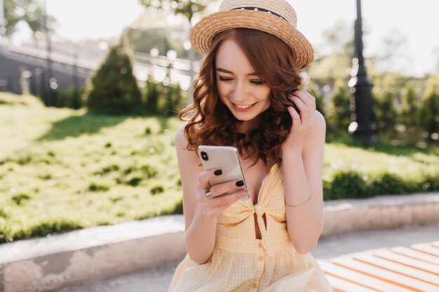 Opgewonden gembermeisje in hoed SMS-bericht met glimlach. Outdoor Portret van sensuele roodharige dame poseren met haar smartphone in park.