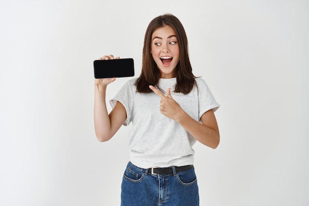 Opgewonden glimlachende vrouw die een smartphone vasthoudt die een leeg scherm van de mobiele telefoon toont en naar de telefoon wijst die op een witte achtergrond staat