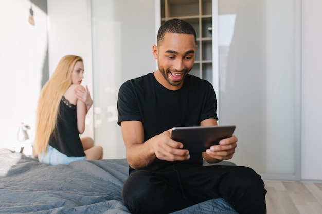 Opgewonden glimlachte knappe jongen spelen op tablet op bed in modern appartement. Boos jonge mooie vrouw met lang blond haar. Plezier hebben, een relatie hebben, thuis