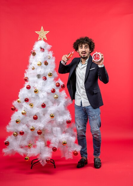 Opgewonden grappige jonge volwassene klok en een glas wijn te houden en permanent in de buurt van de kerstboom op rode stock foto