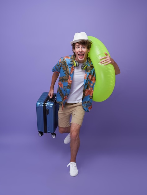 Opgewonden haast jonge man toerist met bagage en rubberen ring loopt naar vakantie op paarse achtergrond