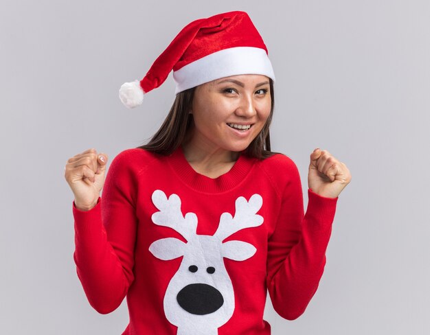 Opgewonden jong Aziatisch meisje dat Kerstmishoed met sweater draagt die ja gebaar toont dat op witte achtergrond wordt geïsoleerd