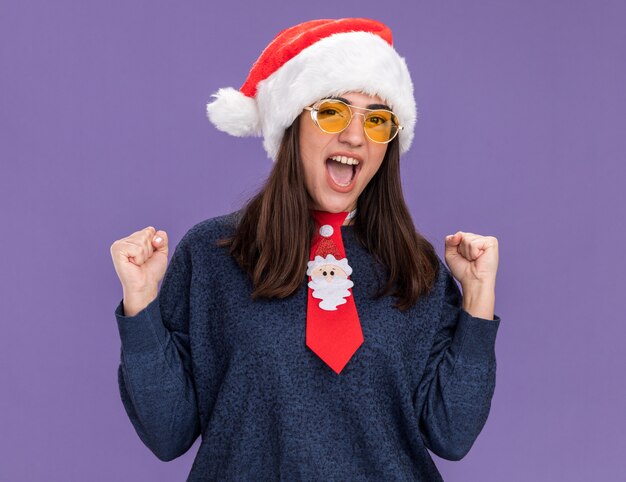 Opgewonden jong kaukasisch meisje in zonnebril met kerstmuts en kerststropdas houdt vuisten geïsoleerd op paarse muur met kopieerruimte