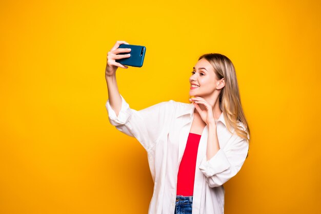 Opgewonden jong meisje dat vrijetijdskleding draagt die zich geïsoleerd over gele muur bevindt, selfie met uitgestrekte hand neemt