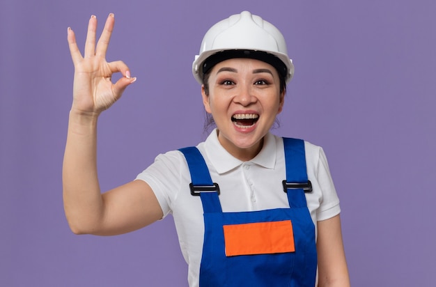 Opgewonden jonge Aziatische bouwer vrouw met witte veiligheidshelm gebaren ok teken