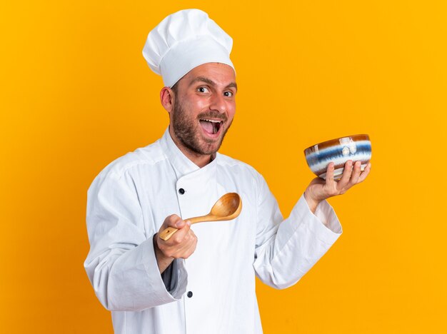 Opgewonden jonge blanke mannelijke kok in uniform van de chef-kok en pet kijkend naar camera met kom en lepel geïsoleerd op een oranje muur