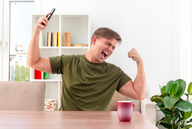 Opgewonden jonge blonde knappe man zit aan tafel met cup tijden biceps en houdt telefoon in de woonkamer