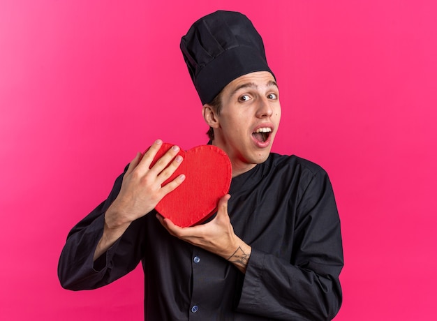 Opgewonden jonge blonde mannelijke kok in chef-kok uniform en pet met hartvorm met beide handen