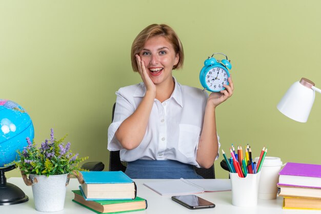 Opgewonden jonge blonde student meisje zit aan bureau met school tools kijken camera hand op gezicht houden wekker geïsoleerd op olijf groene muur