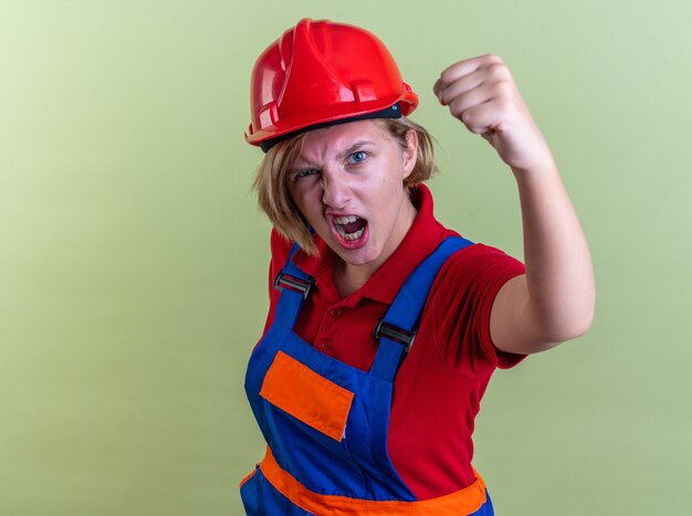 opgewonden jonge bouwer vrouw in uniform met ja gebaar geïsoleerd op olijfgroene muur