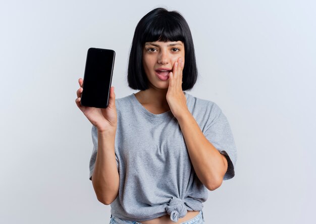 Opgewonden jonge brunette blanke vrouw legt hand op gezicht en houdt telefoon vast