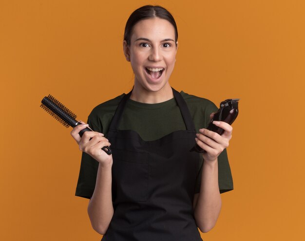 Opgewonden jonge brunette kappersmeisje in uniform houdt tondeuses en kam geïsoleerd op een oranje muur met kopieerruimte