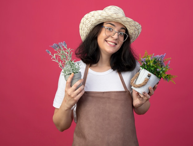 Opgewonden jonge brunette vrouwelijke tuinman in optische bril en uniform dragen tuinieren hoed houdt bloempotten geïsoleerd op roze muur met kopie ruimte