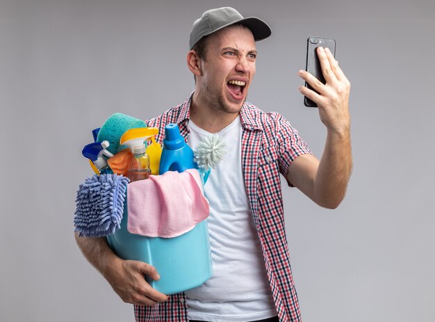 Opgewonden jonge kerel die een dop draagt met een emmer schoonmaakhulpmiddelen spreekt op de telefoon geïsoleerd op een witte achtergrond