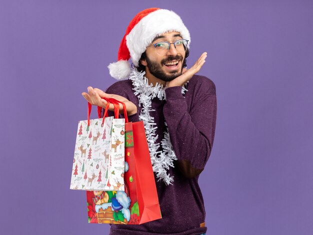 Opgewonden jonge knappe kerel die Kerstmishoed met slinger op hals draagt die giftzakken houdt die handen spreidt die op blauwe muur worden geïsoleerd