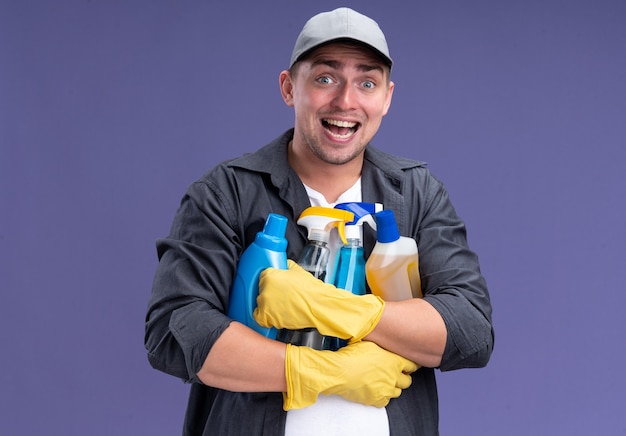 Opgewonden jonge knappe schoonmaakster met t-shirt met pet en handschoenen met schoonmaakgereedschap geïsoleerd op paarse muur
