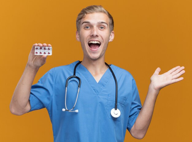 Opgewonden jonge mannelijke arts die artsenuniform met de pillen van de stethoscoopholding draagt die hand verspreiden die op oranje muur wordt geïsoleerd