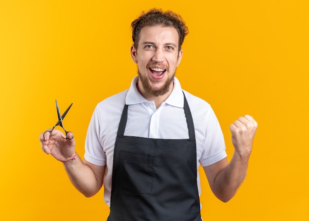 Opgewonden jonge mannelijke kapper die een uniforme schaar draagt met een ja-gebaar dat op een gele muur wordt geïsoleerd