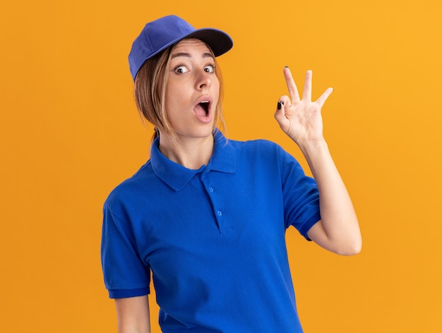 Gratis foto opgewonden jonge mooie bezorgvrouw in uniforme gebaren ok handteken geïsoleerd op oranje muur