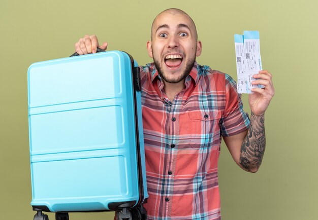 opgewonden jonge reiziger man met koffer en vliegtickets geïsoleerd op olijfgroene muur met kopieerruimte