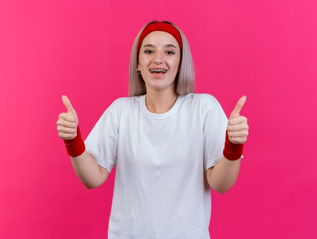 Opgewonden jonge sportieve vrouw met beugels die hoofdband en polsbandjes dragen thumbs up van twee handen geïsoleerd op roze muur