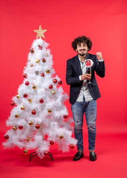 Opgewonden jonge volwassene klok en een glas wijn te houden en permanent in de buurt van de kerstboom op rode stock foto