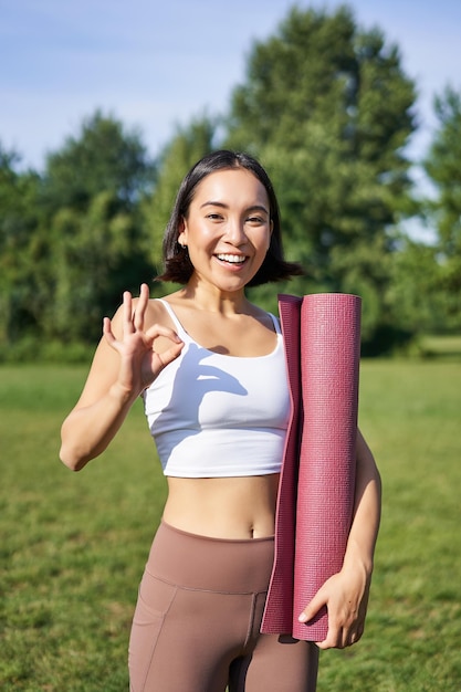 Opgewonden jonge vrouw die met sportmat yogakleding staat, toont een goede tekentraining in parkwelzijn