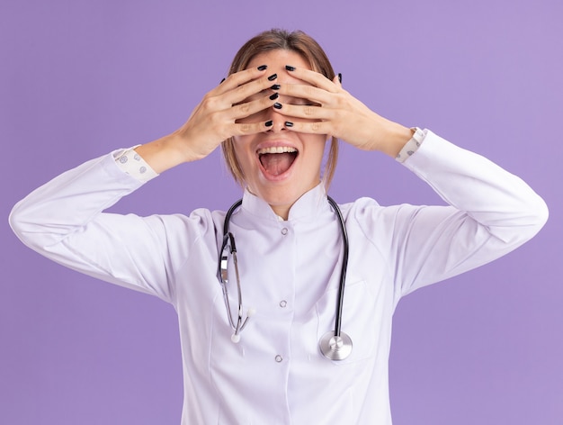 Opgewonden jonge vrouwelijke arts die een medisch gewaad draagt met een stethoscoop bedekte ogen met handen geïsoleerd op een paarse muur