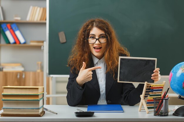 opgewonden jonge vrouwelijke leraar met een bril die vasthoudt en wijst naar een mini-schoolbord dat aan een bureau zit met schoolhulpmiddelen in de klas