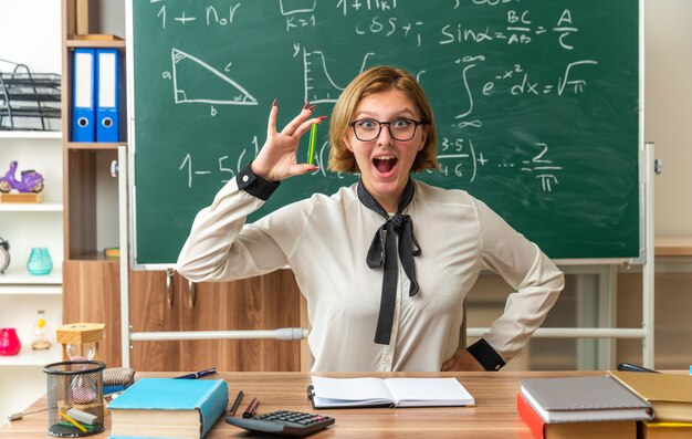 Opgewonden jonge vrouwelijke leraar zit aan tafel met schoolhulpmiddelen die een potlood vasthouden en de hand in de klas zetten