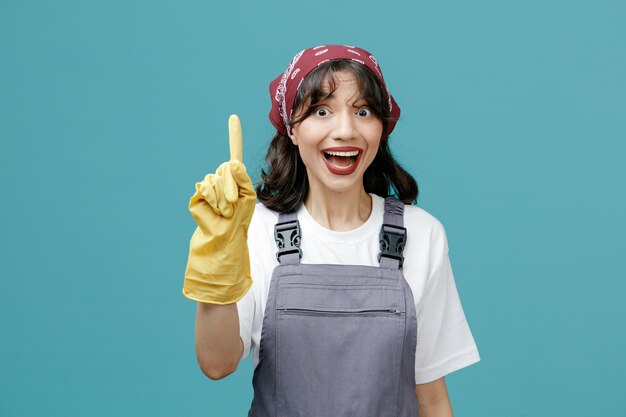 Opgewonden jonge vrouwelijke schoonmaker met een uniforme bandana en rubberen handschoenen die naar de camera kijkt en omhoog wijst geïsoleerd op blauwe achtergrond