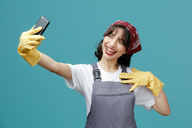Opgewonden jonge vrouwelijke schoonmaker met uniforme bandana en rubberen handschoenen die mobiele telefoon uitrekken en de borst aanraken die selfie geïsoleerd op blauwe achtergrond
