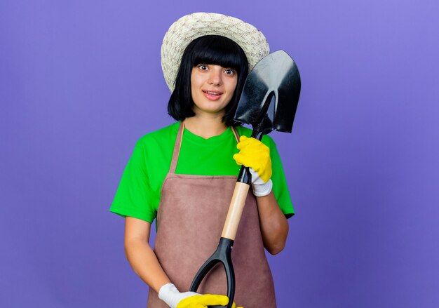 Opgewonden jonge vrouwelijke tuinman in uniform met tuinhoed en handschoenen houdt schop vast