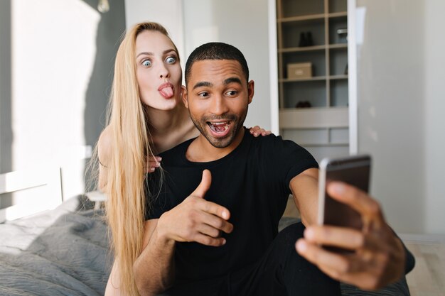 Opgewonden joufyl paar chillen op bed, plezier maken, selfie maken op de telefoon. Goedemorgen samen, tong tonen, relatie, echte positieve emoties uiten, opgewekte stemming