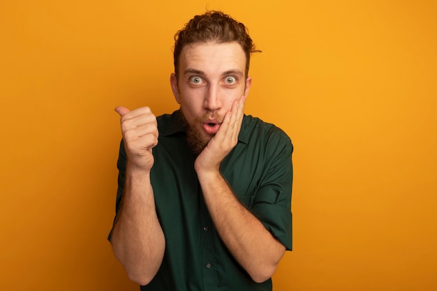 Opgewonden knappe blonde man legt hand op gezicht en wijst terug geïsoleerd op oranje muur