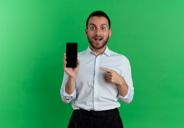 Opgewonden knappe man houdt en wijst op telefoon geïsoleerd op groene muur