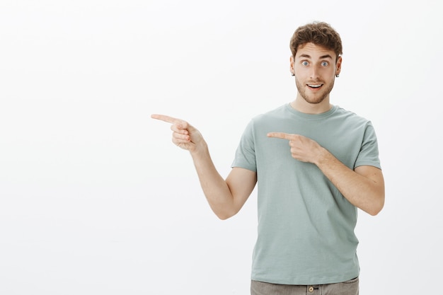 Opgewonden knappe man in oorbellen en t-shirt, naar links wijzend met wijsvingers en breed glimlachend, met de vraag of vrienden met hem mee willen gaan