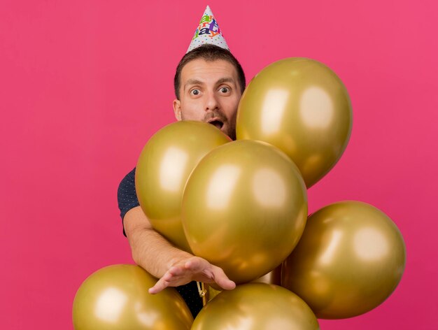 Opgewonden knappe man met verjaardag GLB staat achter helium ballonnen uitrekken hand geïsoleerd op roze muur met kopie ruimte