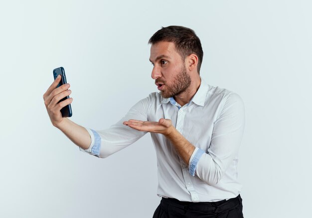 Opgewonden knappe man stuurt kus met hand houden en kijken naar telefoon geïsoleerd op een witte muur