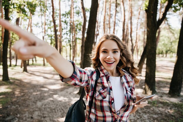 Opgewonden krullend meisje op zoek naar de juiste manier tijdens het wandelen in het bos. Openluchtportret van emotionele vrouwelijke toerist met rugzak.