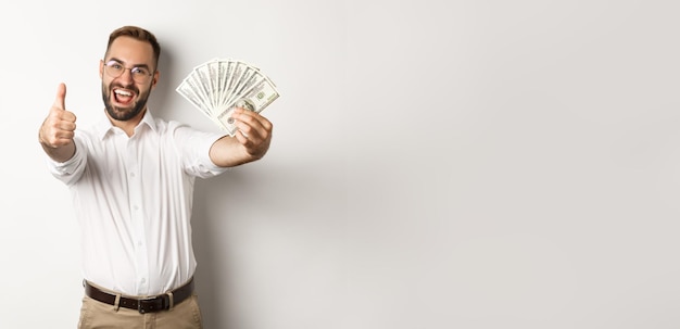 Gratis foto opgewonden man duimen opdagen en geld verdienen geld permanent op witte achtergrond