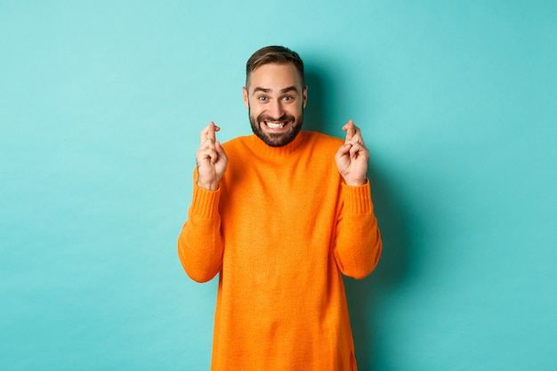 Opgewonden man met baard, die een wens doet, de vingers gekruist houdt voor geluk en glimlacht, staande over de lichtblauwe muur.