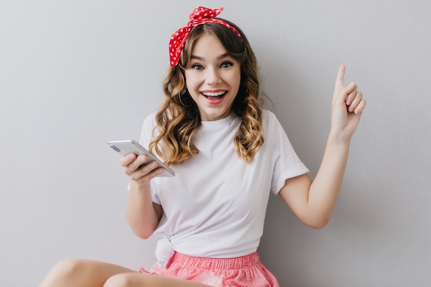 Opgewonden meisje met schattig rood lint poseren met telefoon. Sensuele jonge dame die haar apparaat vasthoudt en geluk uitdrukt.