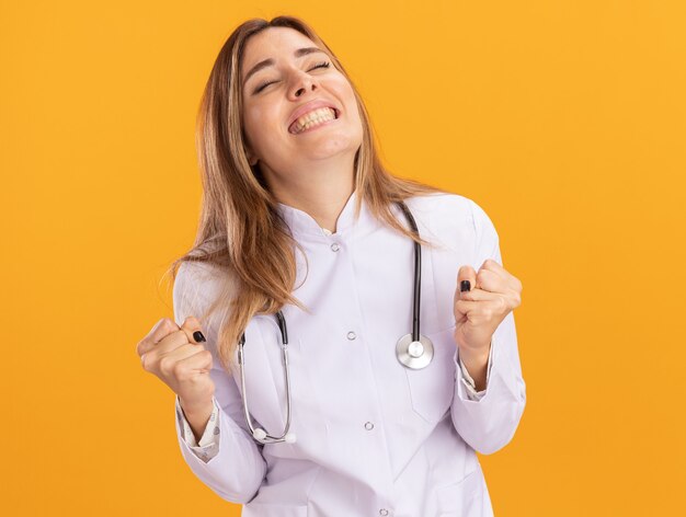 Opgewonden met gesloten ogen jonge vrouwelijke arts die medische robe met stethoscoop draagt die ja gebaar toont dat op gele muur wordt geïsoleerd