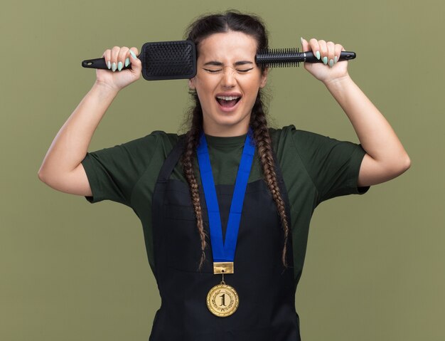 Opgewonden met gesloten ogen jonge vrouwelijke kapper in uniform en medaille houden kammen op tempels geïsoleerd op olijfgroene muur