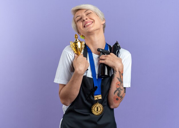 Opgewonden met gesloten ogen kantelend hoofd jonge mooie vrouwelijke kapper in uniform dragen medaille bedrijf kapper tools en winnaar beker geïsoleerd op blauwe muur