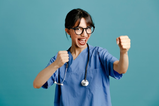 Opgewonden met sterk gebaar jonge vrouwelijke arts die uniforme stethoscoop draagt die op blauwe achtergrond wordt geïsoleerd